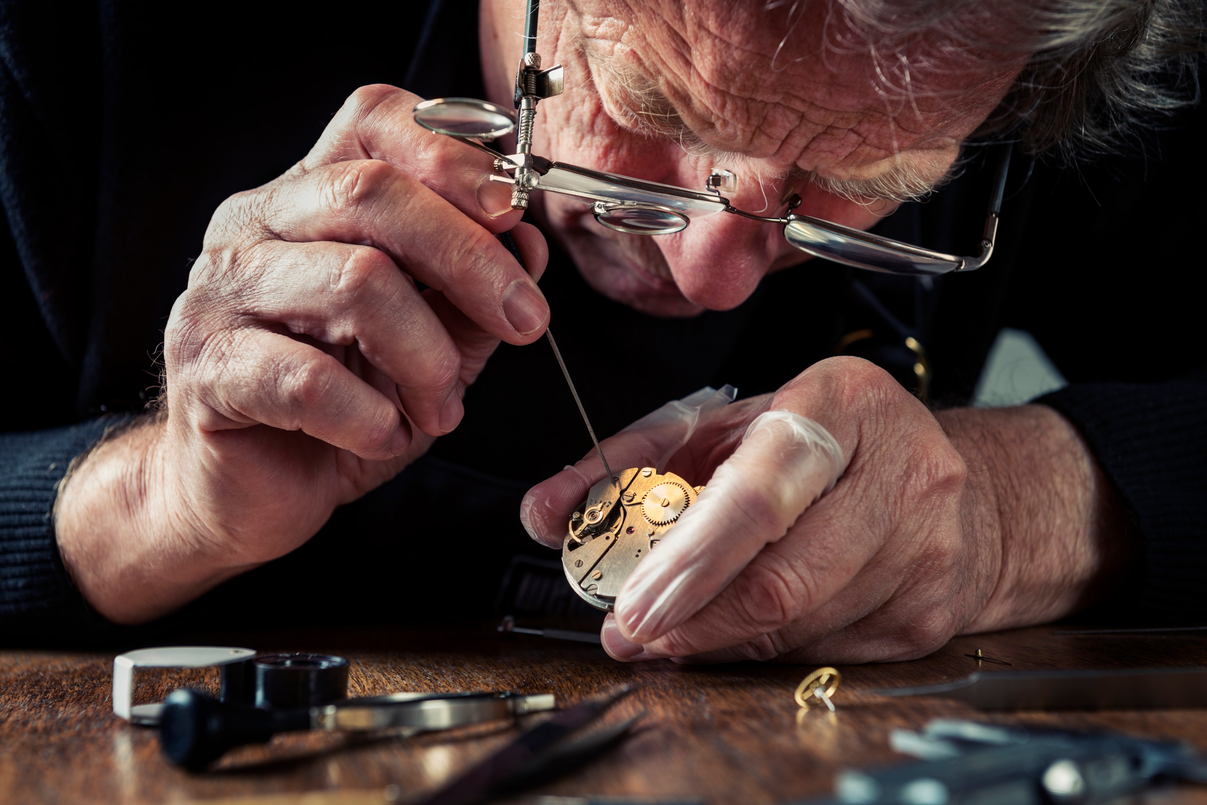 A master horologist examining a watch movement under loupe-glasses at the bench.